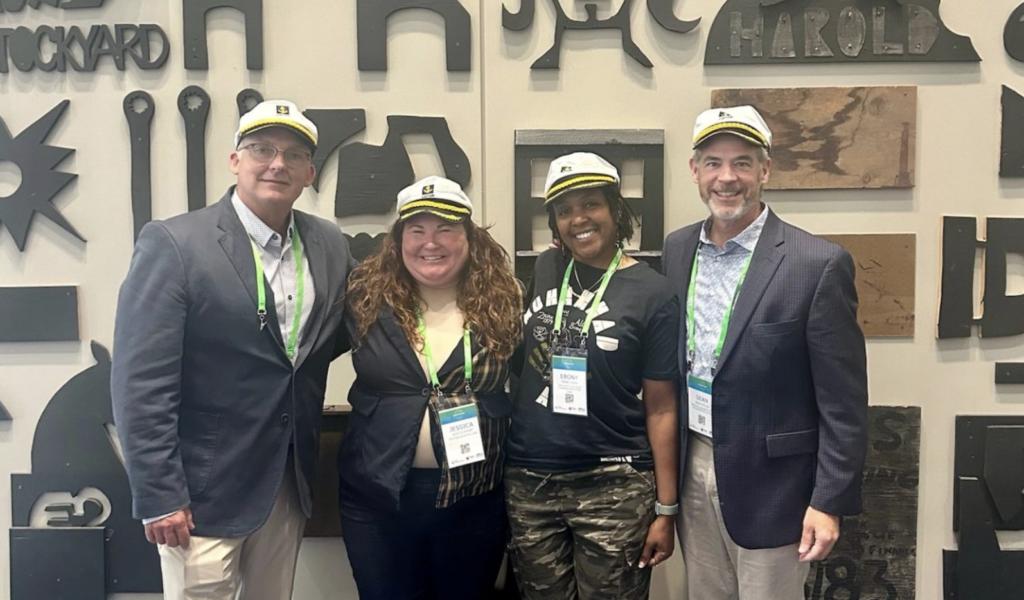 Four convening attendees wearing matching captain-style hats with yellow bands pose together against a wall decorated with various industrial-style letters and designs. The group wears conference lanyards with green badges and a mix of business and casual attire.